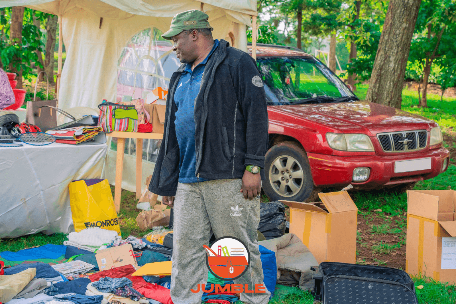 Man at outdoor jumble sale with clothes and boxes, standing near a red car.