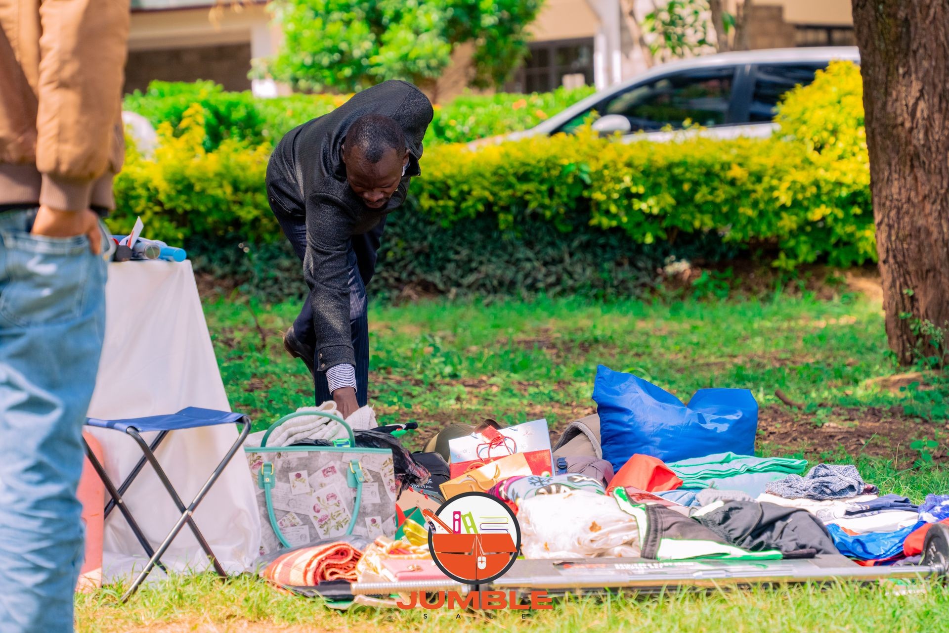 Man setting up a clothes stall outdoors with various items on display at a park.