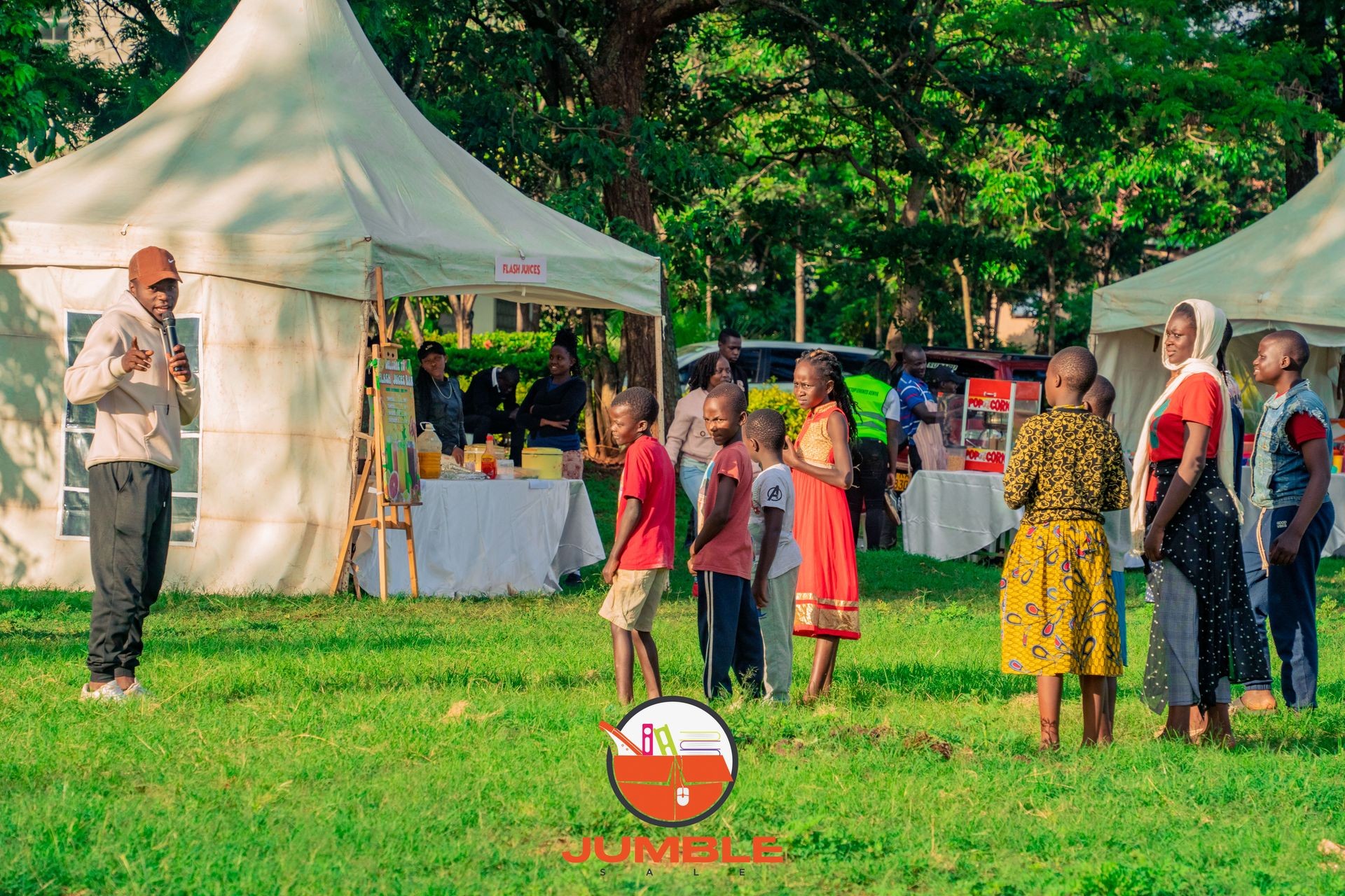 Outdoor event with people gathered near a tent, including children and a speaker holding a microphone.