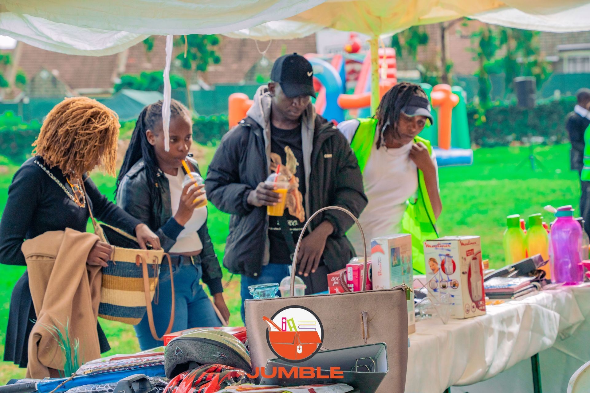 People standing by a table with various products and drinks under a tent at an outdoor event.