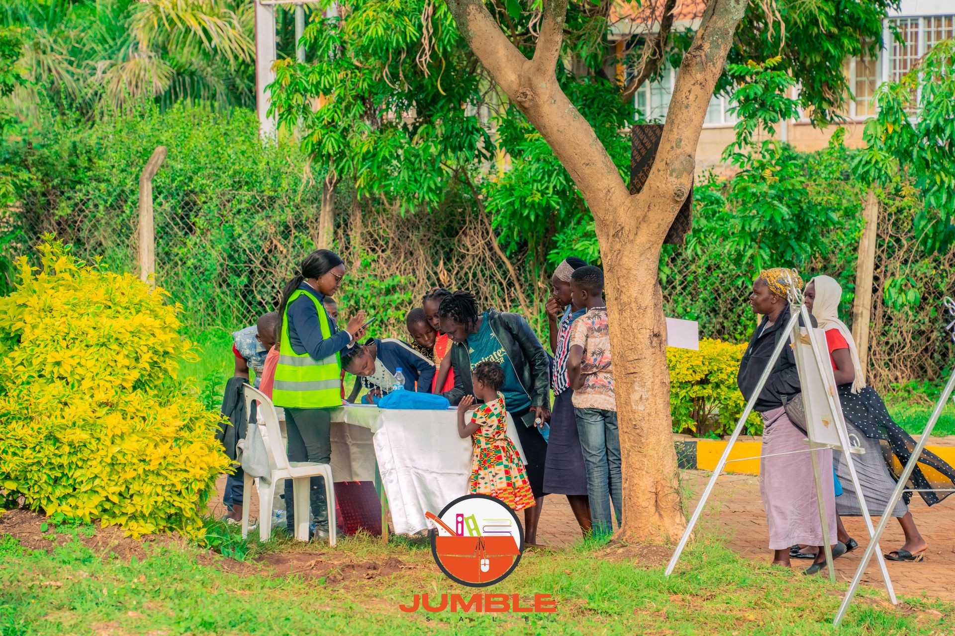 People gathered around a table outdoors, with a person in a reflective vest assisting them.